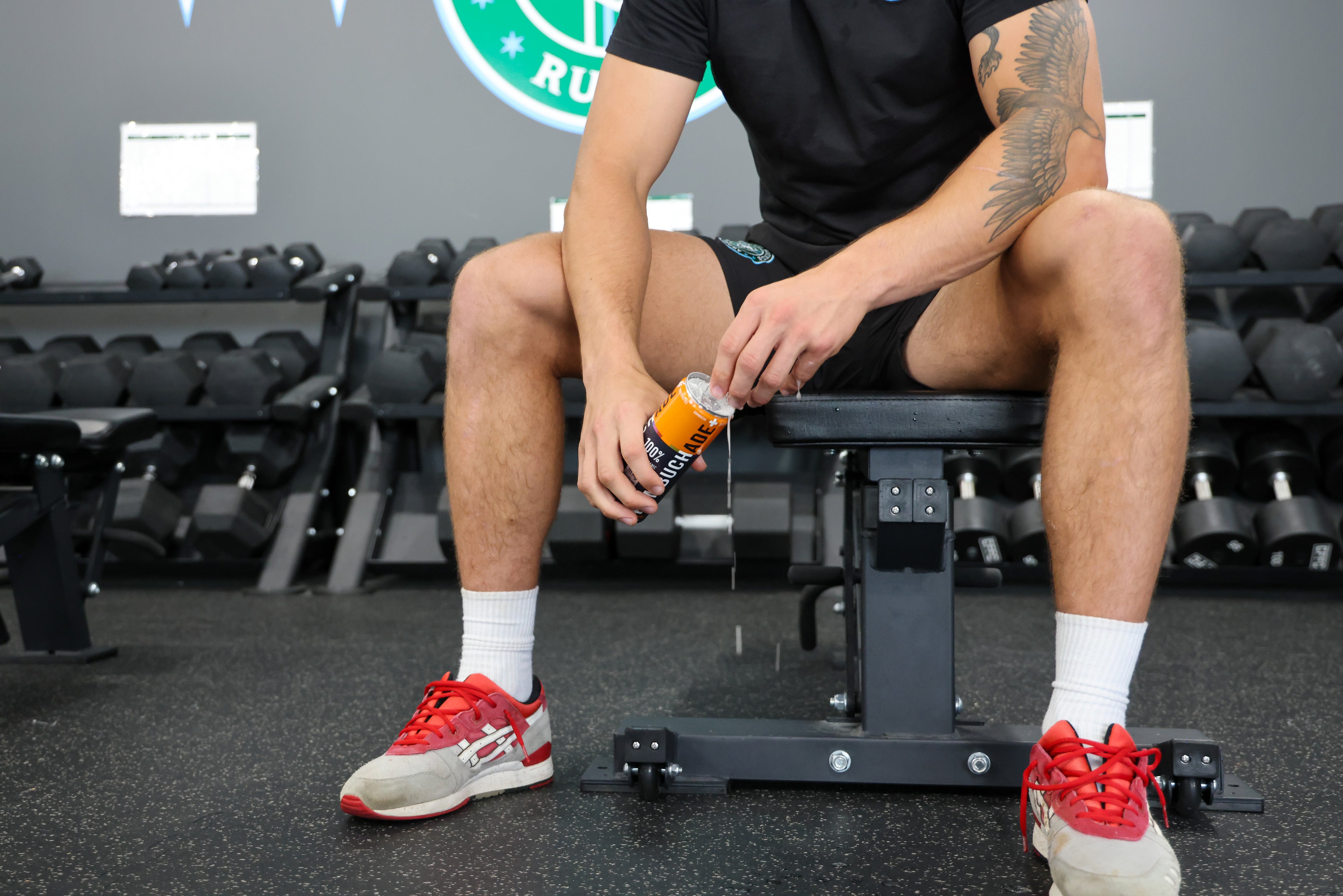 a fitness enthusiast sitting on a bench in front of a weight rack opening a can of Wild Guava Kombuchade