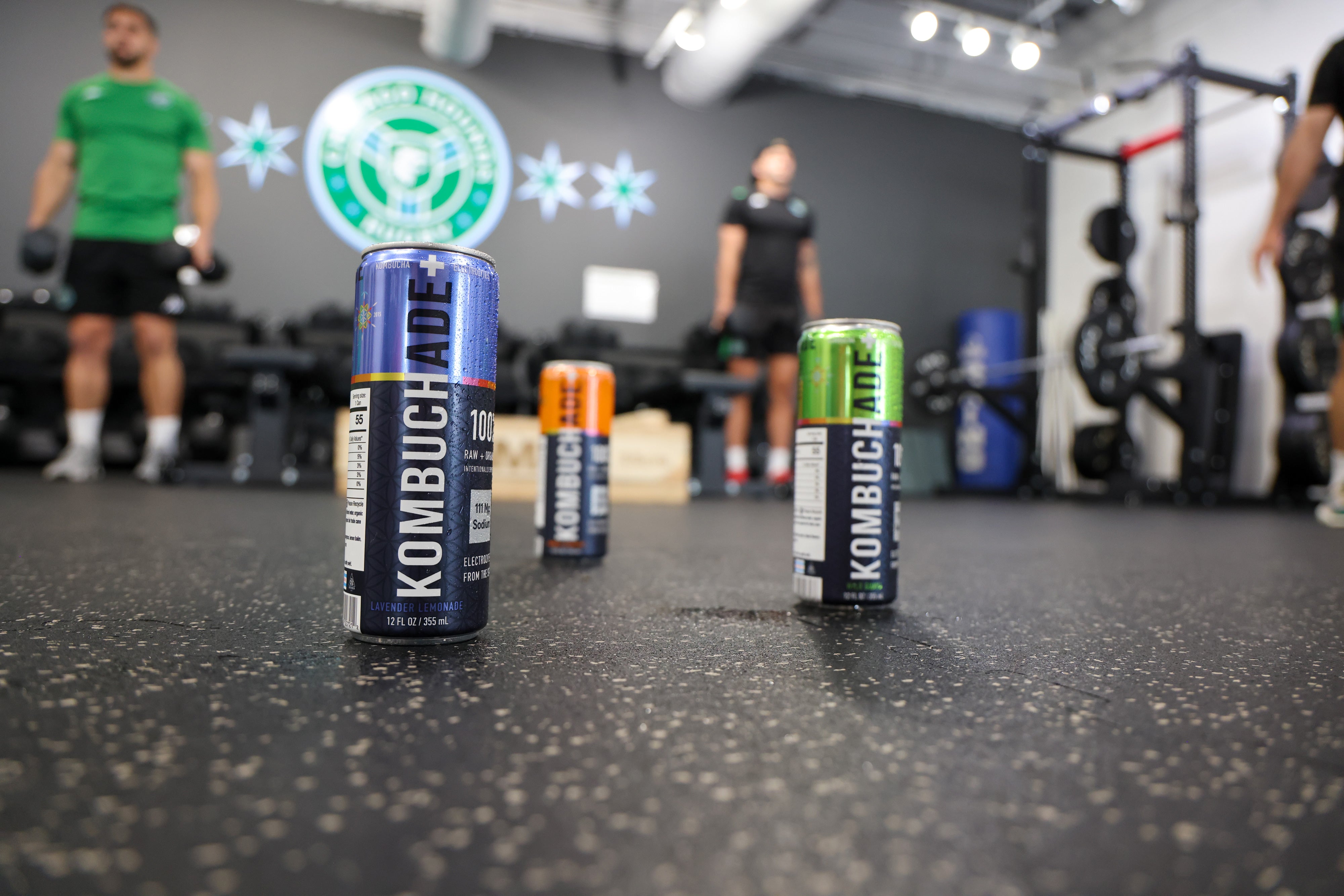 three cans of Kombuchade sitting on the floor of a gym while athletes work out