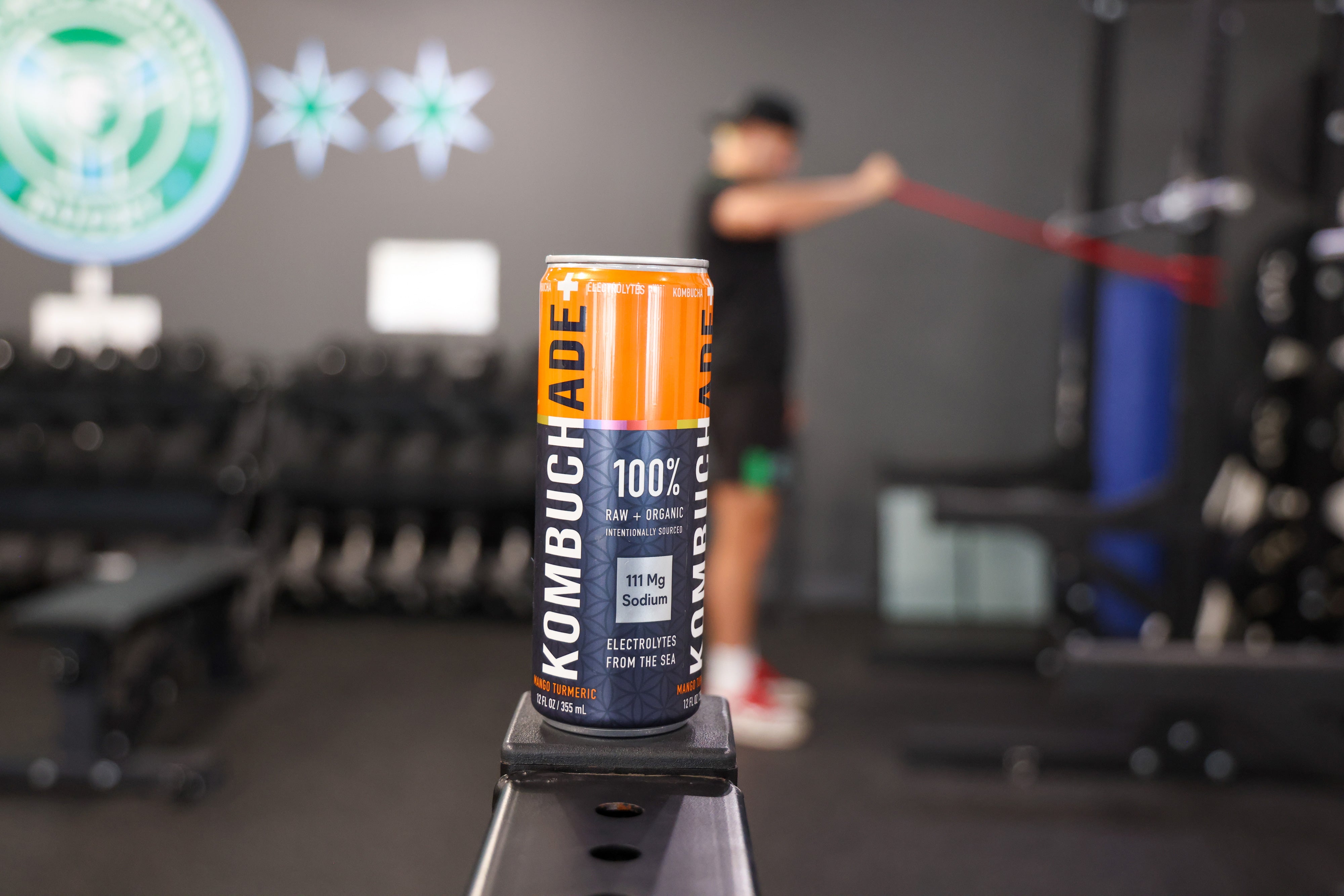 a can of mango turmeric Kombuchade sitting on a piece of gym equipment while an athletes works out