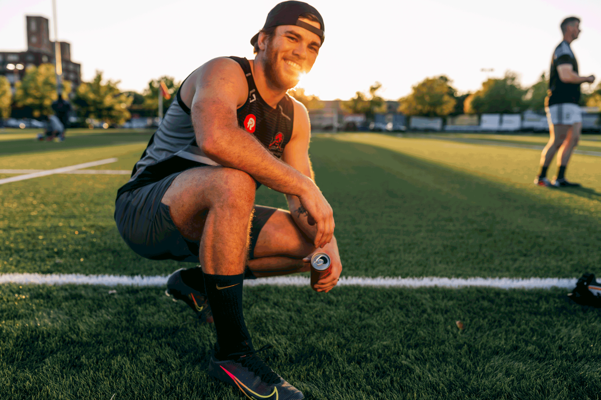 Matt Lancor founder of Kombuchade kneeling with can of Kombuchade on rugby field