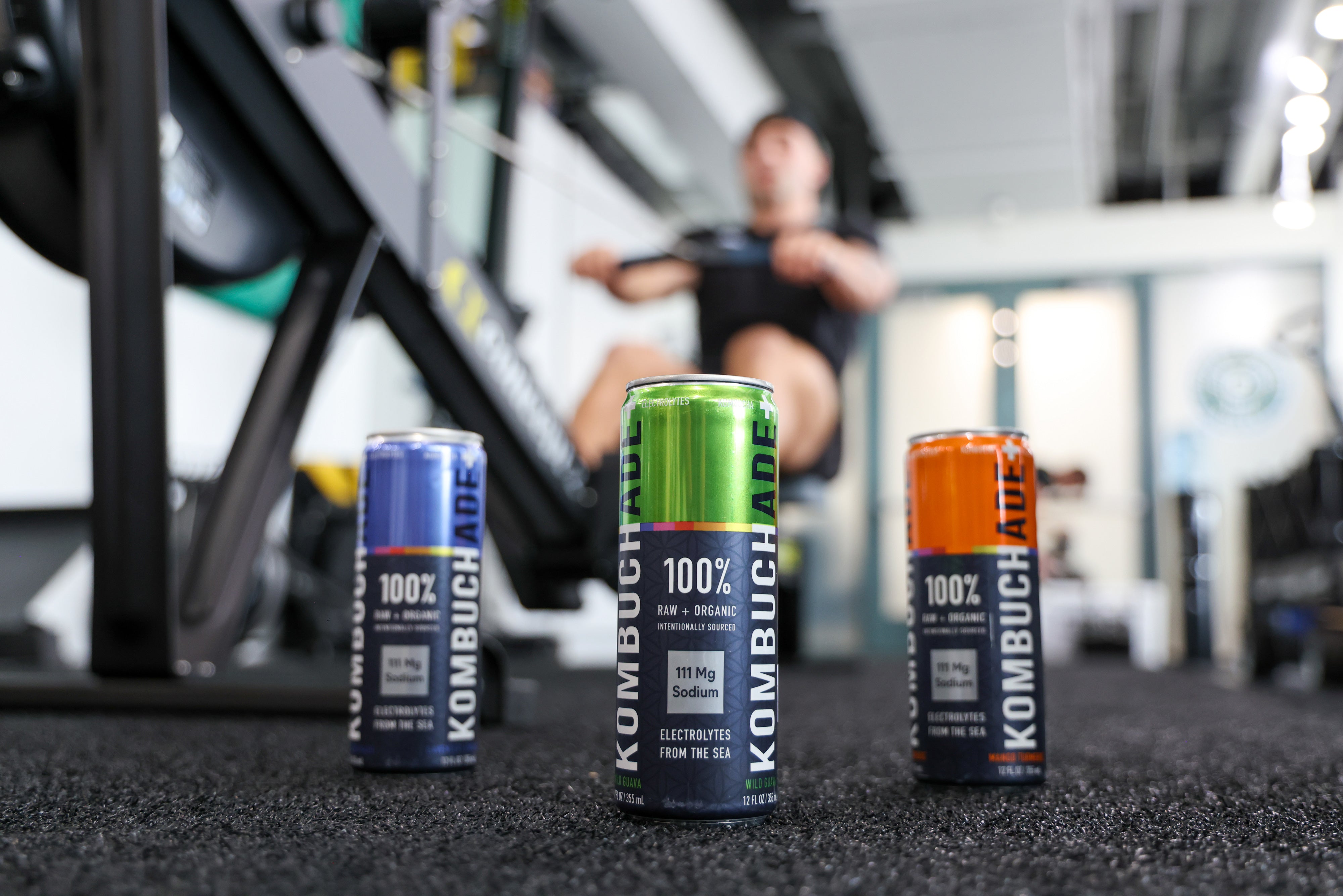 Athlete in gym on a rower with three cans of Kombuchade in all three flavors