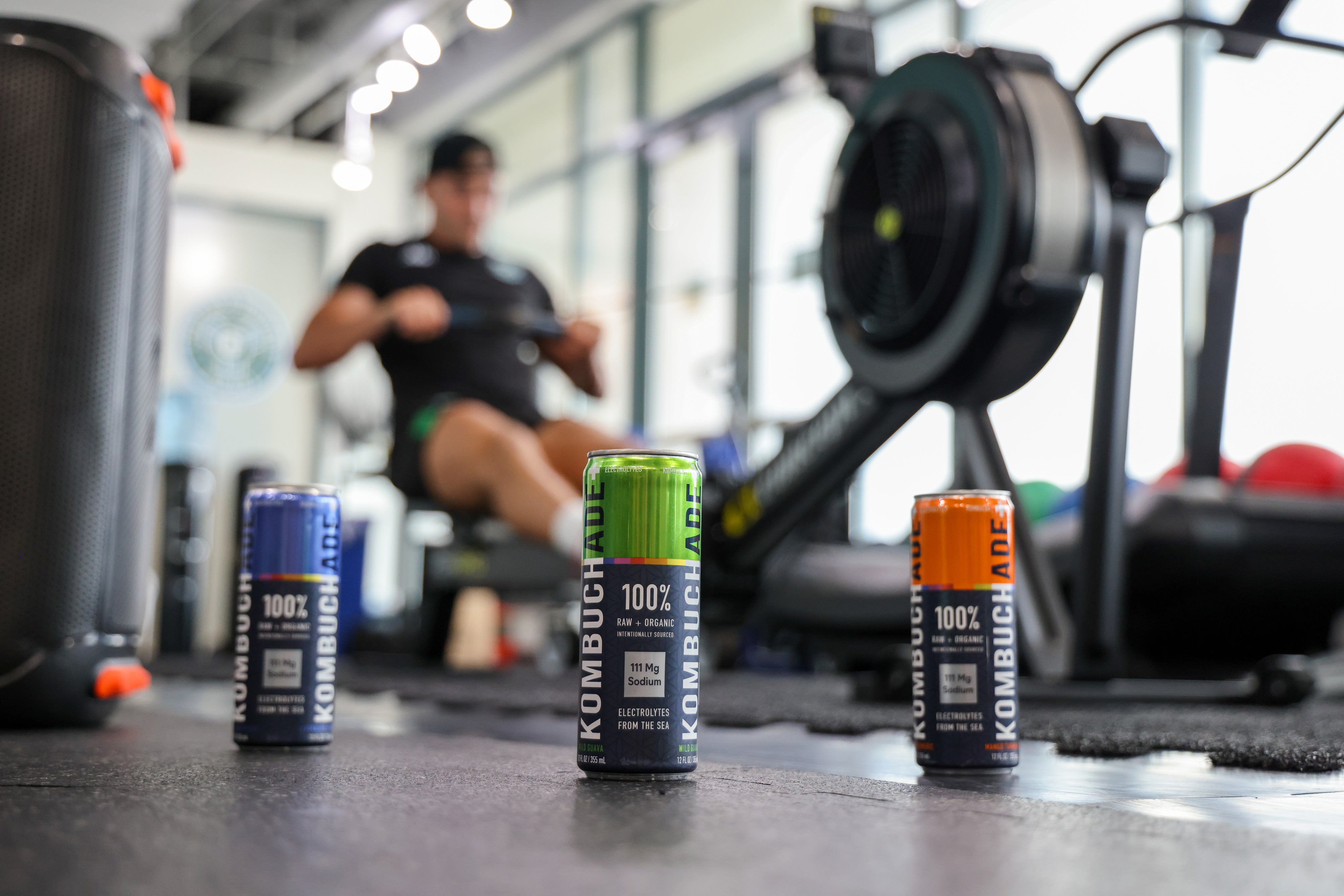 Athlete in gym on a rower with three cans of Kombuchade in all three flavors